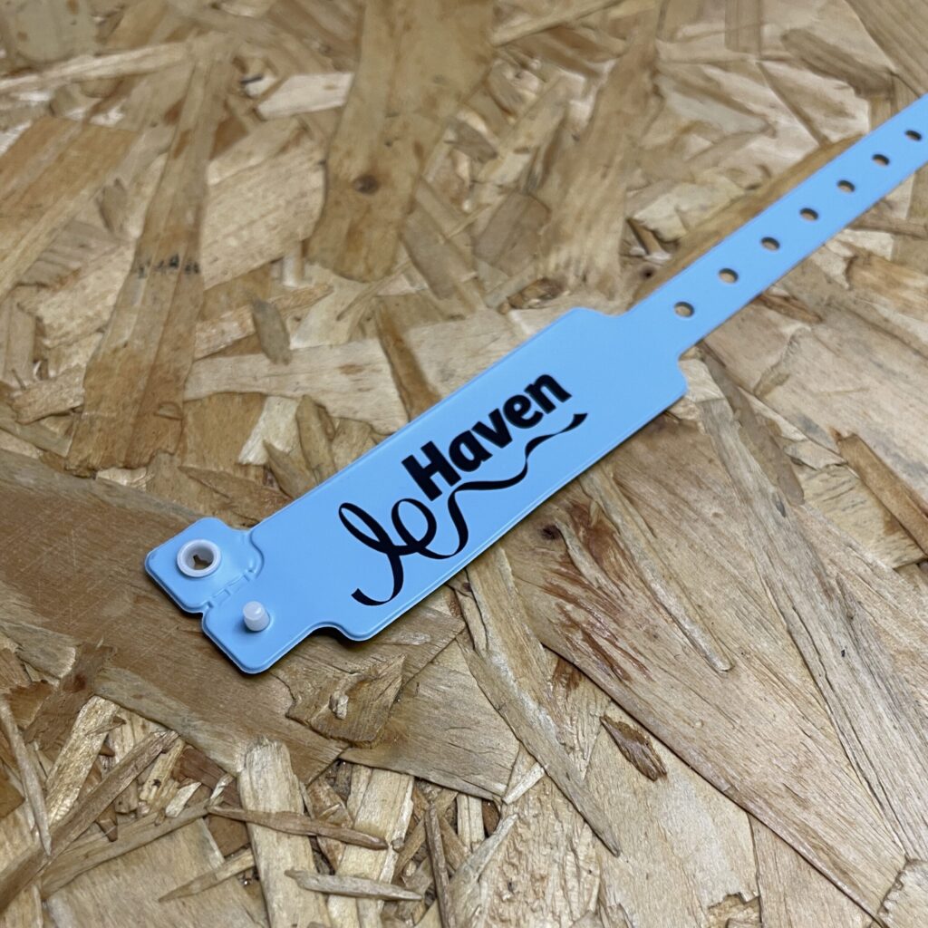 A single light blue wide-face vinyl event wristband featuring a custom black "Haven" logo and a white plastic locking snap closure on a textured wood particle board background.