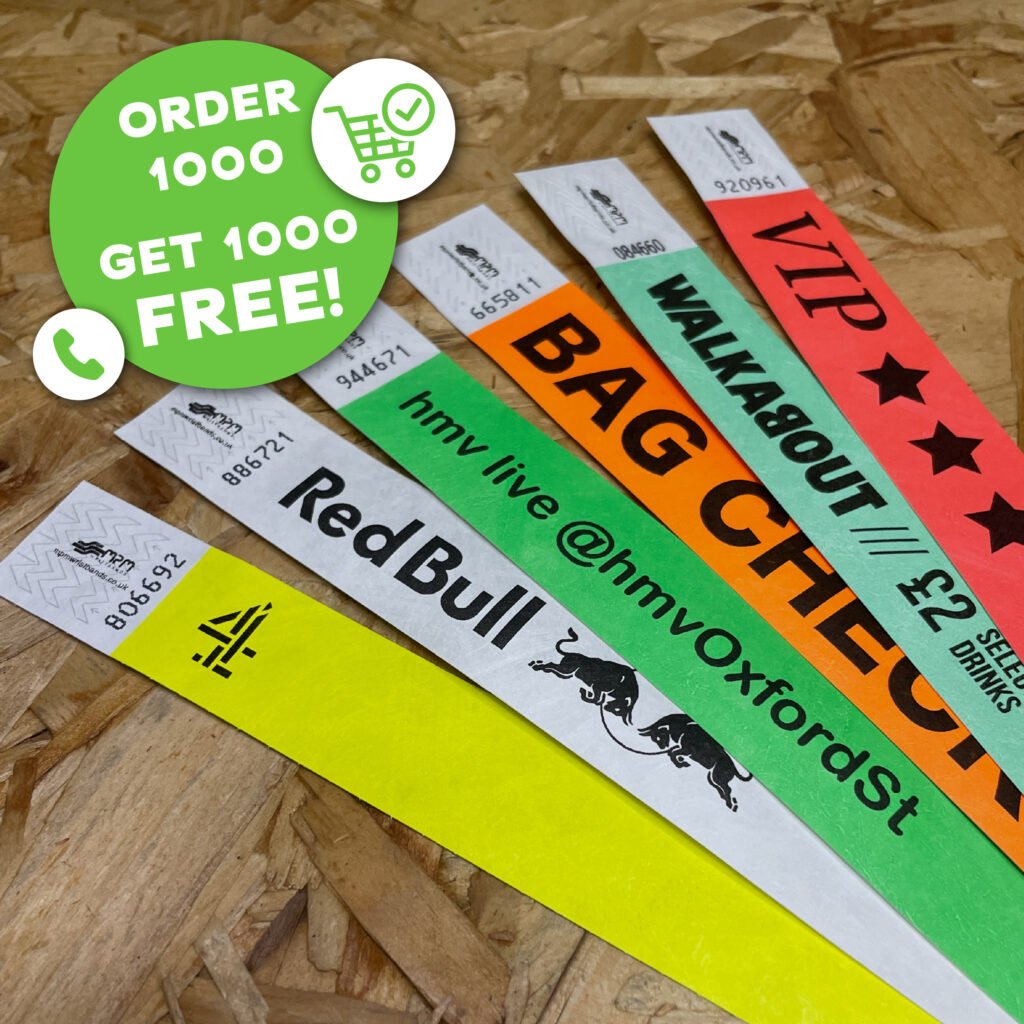 Five colorful Tyvek wristbands with custom logos like Red Bull and Channel 4 on a wood background with a "Buy 1000 Get 1000 Free" badge.