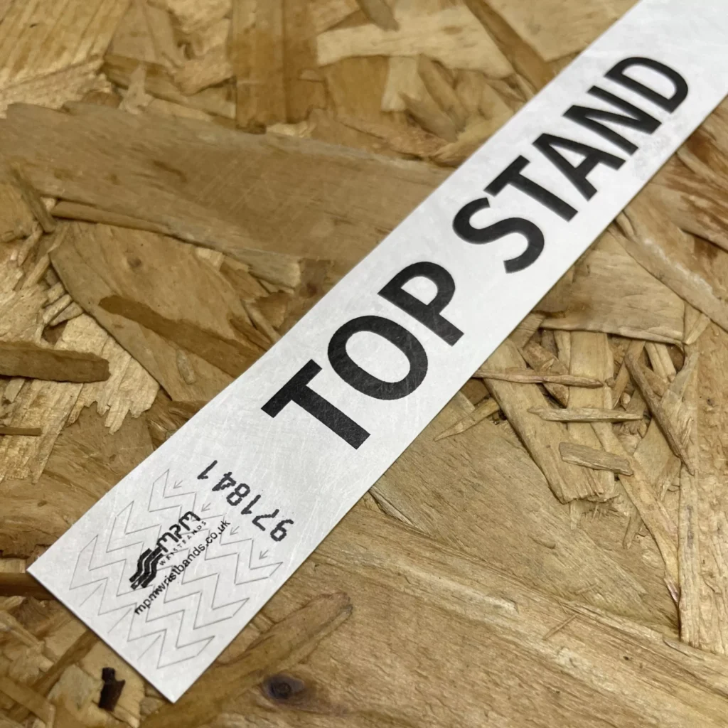 A white Tyvek wristband featuring bold black "TOP STAND" text and sequential numbering on a wood surface.
